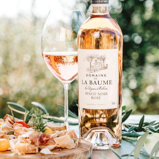 Bottle of Domaine de La Baume Rosé wine with a glass and appetizers on a table outdoors.